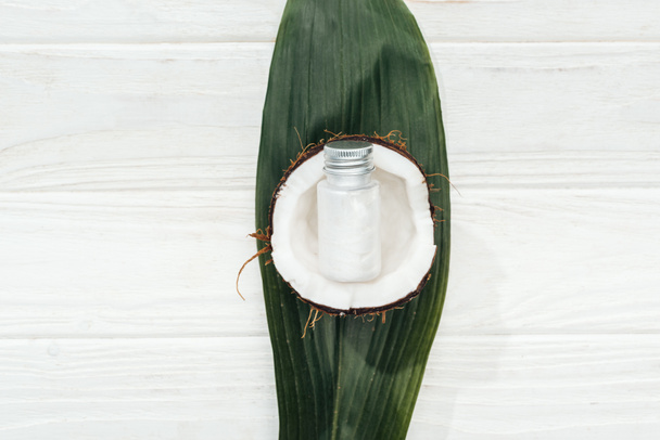 Draufsicht auf Kokosnuss-Schönheitsprodukt in Flasche auf Kokosnuss-Hälfte und grünem Palmblatt auf weißer Holzoberfläche - Foto, Bild