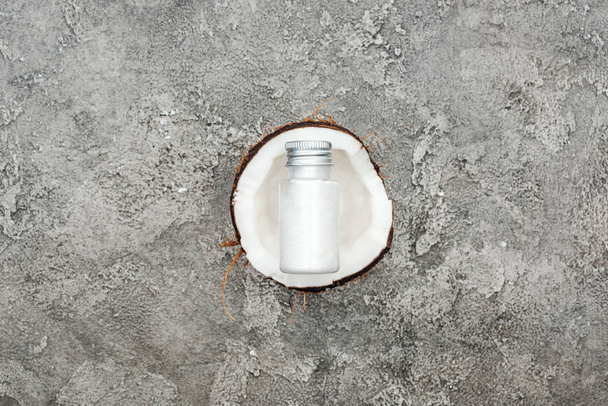 Draufsicht der Creme in der Flasche auf Kokosnusshälfte auf grauem strukturiertem Hintergrund - Foto, Bild