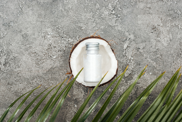 Top uitzicht van crème in fles op kokosnoot half op grijs getextureerde achtergrond met Palm blad - Foto, afbeelding