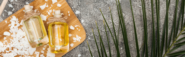 вид сверху на кокосовое масло в бутылках на деревянной доске на сером текстурированном фоне с пальмовым листом, панорамный снимок
 - Фото, изображение