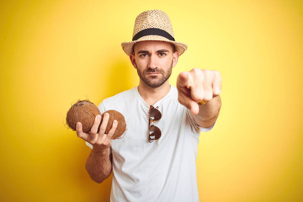 Mladý muž, který nosí Letní klobouk a drží kokosový plod na žluté pozadí ukazující prstem na kameru a na vás, znamení ruky, pozitivní a sebevědomé gesto zepředu - Fotografie, Obrázek
