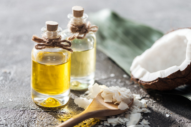 selective focus of bottles with coconut oil near coconut half and shavings in wooden spoon on grey background - Φωτογραφία, εικόνα
