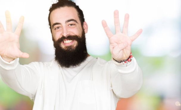 Молодой человек с длинными волосами и бородой в спортивной толстовке показывает и показывает пальцами номер девять улыбаясь уверенно и счастливо
. - Фото, изображение