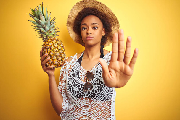 Fiatal afro-amerikai nő afro hajú gazdaság pineaple több mint sárga elszigetelt háttér nyitott kézzel csinál stop jel komoly és magabiztos kifejezés, védelmi gesztus - Fotó, kép