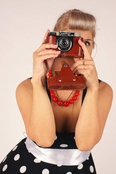 Beautiful blond woman in pinup style, dressed in a polka dot dress keeps the camera in front of her, taking a picture, white background - Foto, Bild