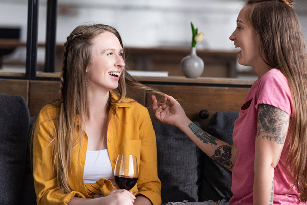 uśmiechnięty lesbijek trzymając kieliszek do wina i patrząc na dziewczynę, siedząc na kanapie w salonie - Zdjęcie, obraz