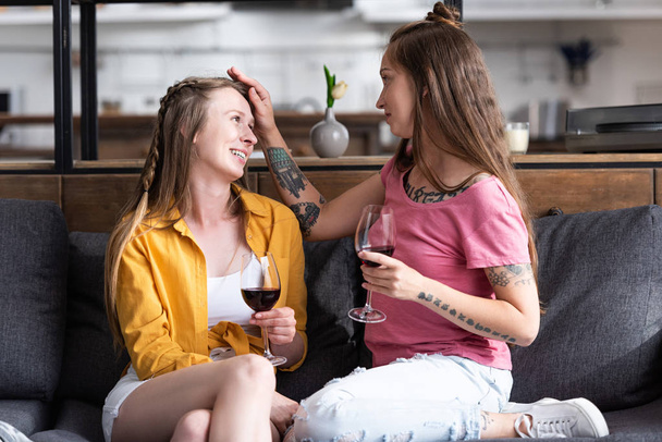 two lesbians holding wine glasses and looking at each other while sitting on sofa in living room - Photo, Image