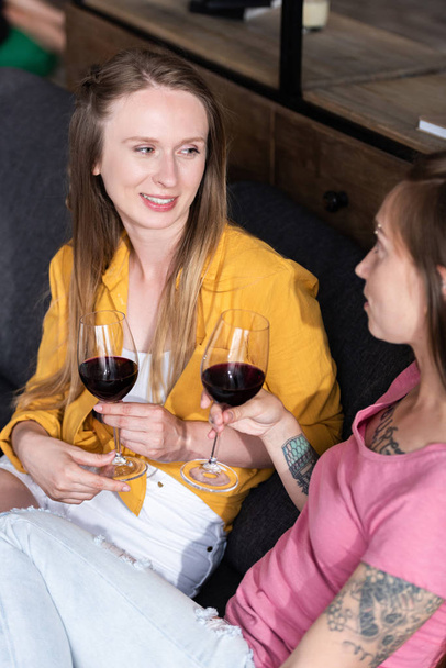 dva lesbičky držící vinnou skleničky a seděli na pohovce v obývacím pokoji - Fotografie, Obrázek