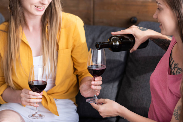 partial view of two lesbians holding wine glasses and pouring wine while sitting on sofa in living room - Fotó, kép