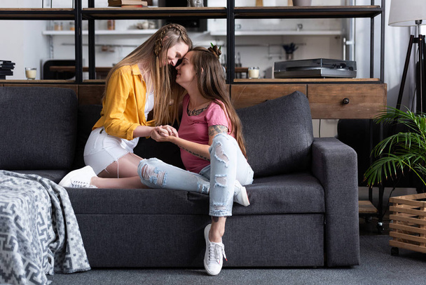 two smiling lesbians holding hands while sitting on sofa in living room - Foto, Imagem
