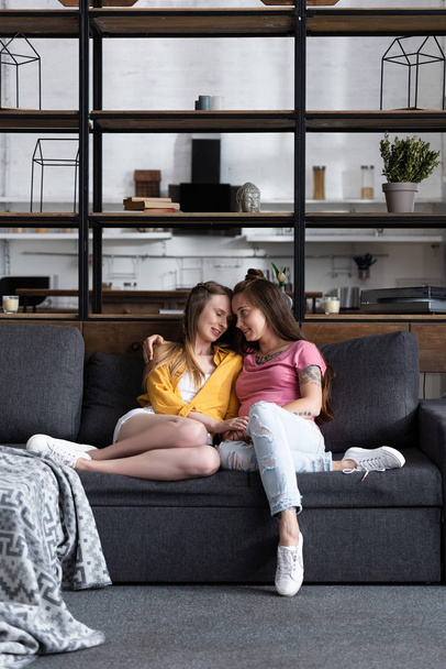 two pretty lesbians embracing while sitting on sofa in living room - Photo, Image