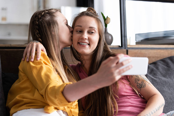 lesbian in yellow blouse kissing girlfriend while taking selfie - Photo, Image