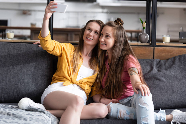 δύο χαμογελαστά κορίτσια κάθονται στον καναπέ και παίρνοντας selfie στο σαλόνι - Φωτογραφία, εικόνα