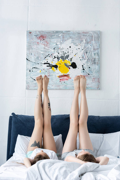 two barefoot lesbians lying on bed with legs up in morning - Фото, изображение