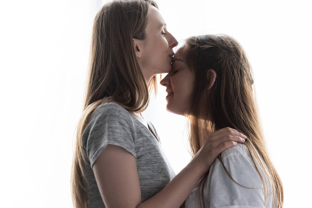 Zijaanzicht van lesbische omarmen vriendin en zoenen haar in voorhoofd thuis - Foto, afbeelding