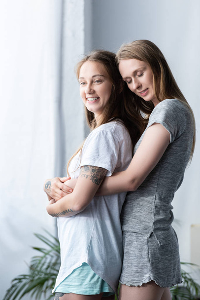 dos lesbianas sonrientes en camisetas abrazándose en el dormitorio
 - Foto, Imagen