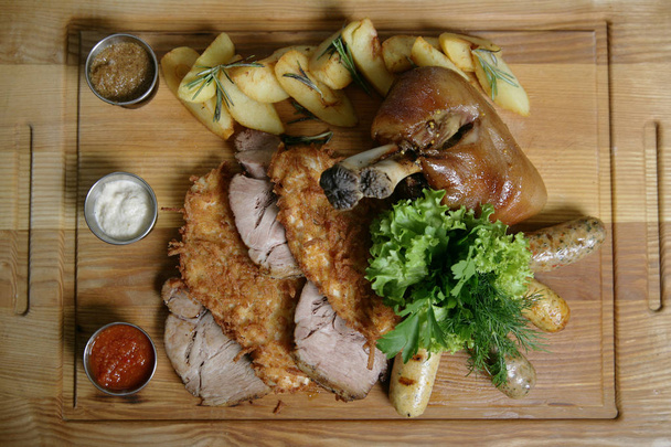 Mix food for a big company. A big delicious whole baked knuckle, fried meat chops, ham, potatoes. With greens and sauces. On a wooden board.  - Photo, Image