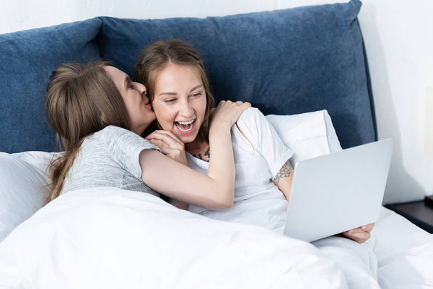 two smiling lesbians embracing and kissing while using laptop in bed in morning - 写真・画像