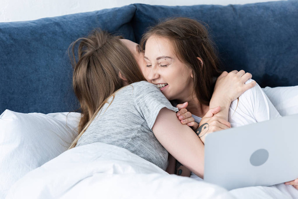 two smiling lesbians embracing while using laptop in bed in morning - Fotoğraf, Görsel