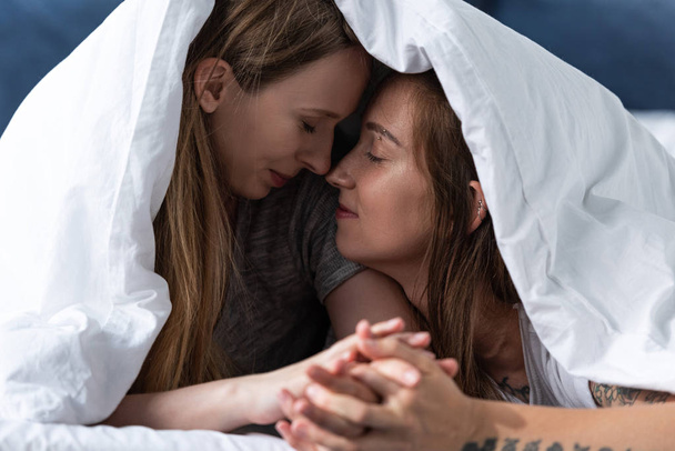 two happy lesbians holding hands while lying under blanket on bed - Photo, Image