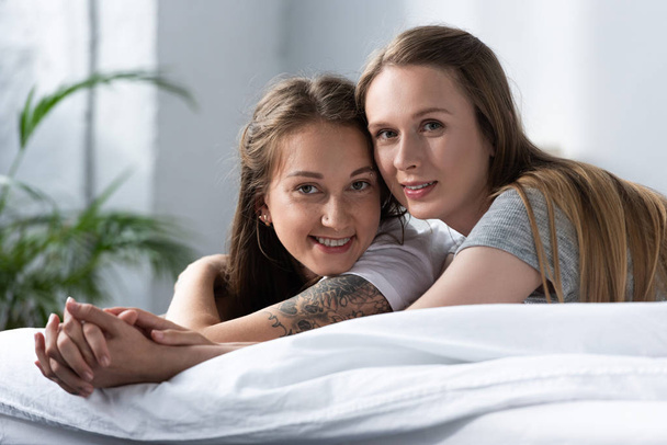 two smiling lesbians holding hands and looking at camera while lying on bed in morning - Photo, Image