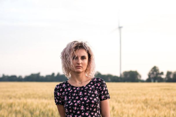 young tanned curly haired woman in dress is happy to be on the field of ripened wheat, wind turbine on the background - Foto, imagen