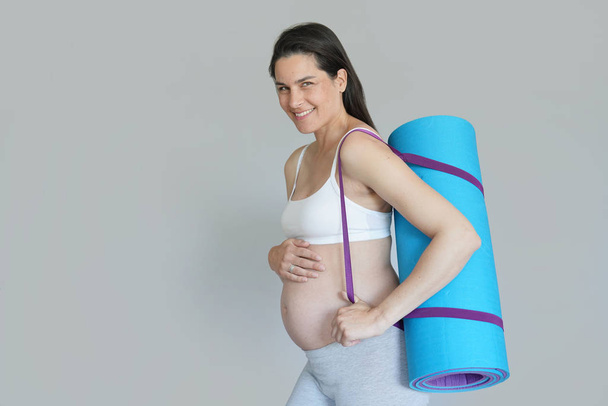 Pregnant woman carrying fitness mat, isolated - Фото, изображение