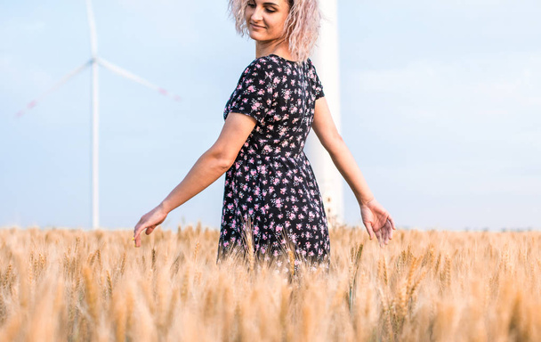 young tanned curly haired woman in dress is happy to be on the field of ripened wheat, wind turbine on the background - Zdjęcie, obraz