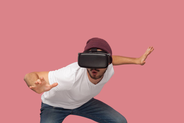 shocked bearded young man in white shirt and casual hat wearing vr headset playing video game with raised arms on pink background - Foto, Imagen