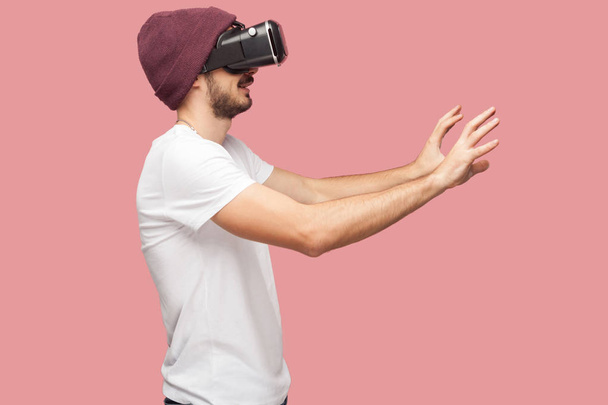 Side view of shocked bearded young man in white shirt and casual hat wearing vr headset playing video game on pink background - Zdjęcie, obraz