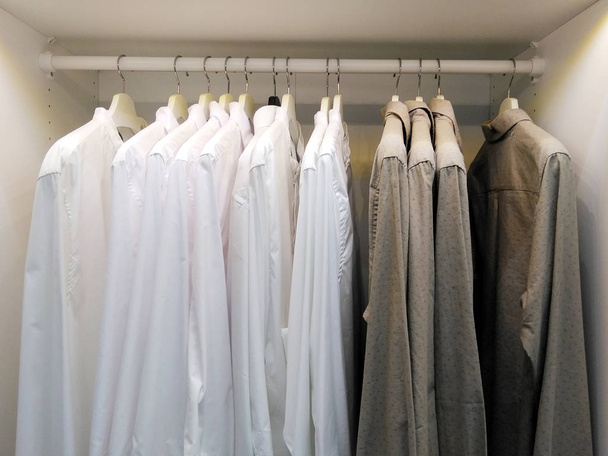 Ряд серых и белых рубашек висит на вешалке в белом шкафу. Современный деревянный гардероб с мужской одеждой висит на рельсах в прогулке в шкафу, в скандинавском стиле
. - Фото, изображение