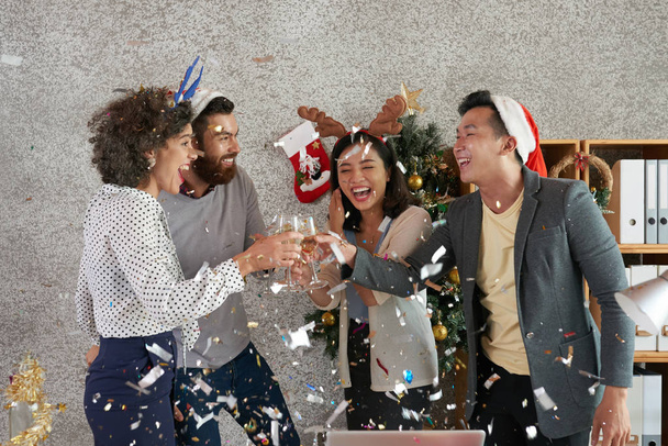 Laughing excited multi-ethnic coworkers toasting with champagne glasses under falling confetti - Photo, Image