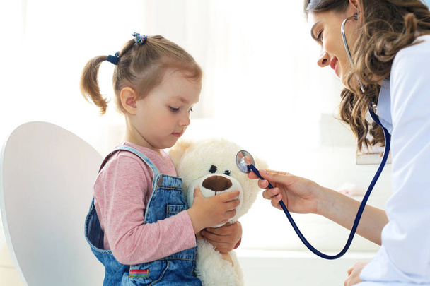 Little girl at the doctor for a checkup. Doctor playfully checking the heart beat of a teddy bear. - Foto, Imagem