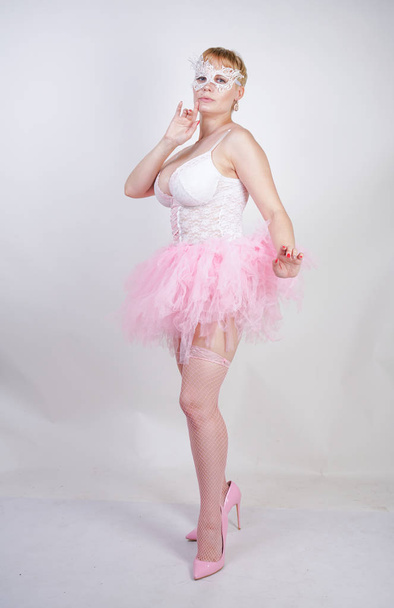 pretty plus size playful adult girl in white lace corset and tutu skirt standing alone on studio background. cute blonde chubby woman in pink dress posing as a doll. - Фото, зображення