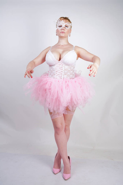 pretty plus size playful adult girl in white lace corset and tutu skirt standing alone on studio background. cute blonde chubby woman in pink dress posing as a doll. - Photo, Image