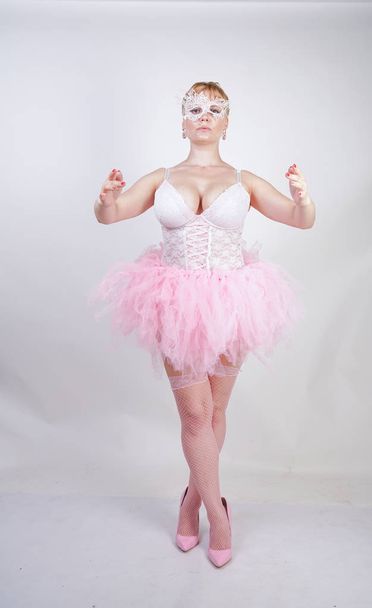 pretty plus size playful adult girl in white lace corset and tutu skirt standing alone on studio background. cute blonde chubby woman in pink dress posing as a doll. - Photo, Image