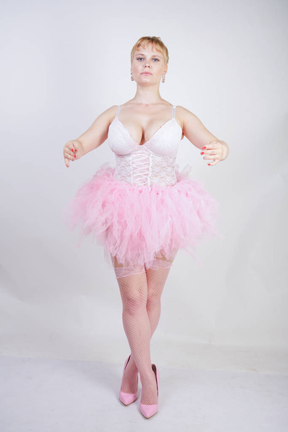 hübsche Plusgröße verspieltes erwachsenes Mädchen in weißem Spitzenkorsett und Tutu-Rock, das allein auf Studiohintergrund steht. süße blonde mollige Frau in rosa Kleid posiert als Puppe. - Foto, Bild