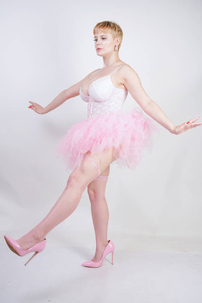 pretty plus size playful adult girl in white lace corset and tutu skirt standing alone on studio background. cute blonde chubby woman in pink dress posing as a doll. - Valokuva, kuva