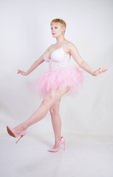 pretty plus size playful adult girl in white lace corset and tutu skirt standing alone on studio background. cute blonde chubby woman in pink dress posing as a doll. - Foto, Imagem