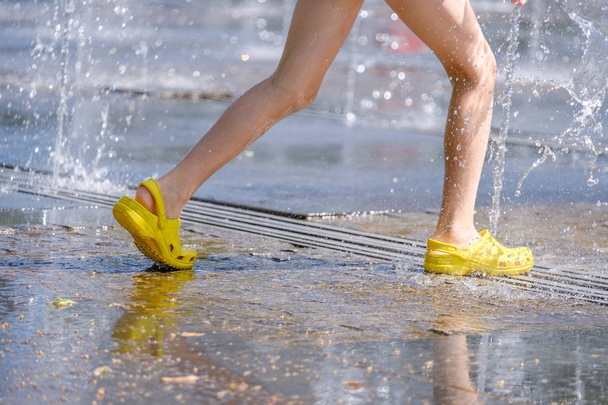 Legs of a child in yellow rubber shoes running through the refreshing splashes of the city fountain on a hot summer day - Photo, image