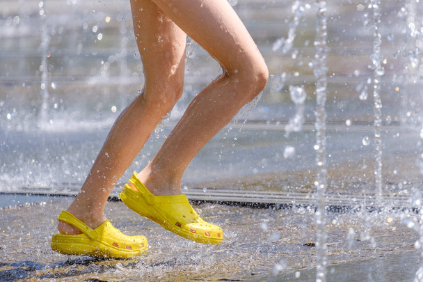 Legs of a child in yellow rubber shoes running through the refreshing splashes of the city fountain on a hot summer day - Photo, image