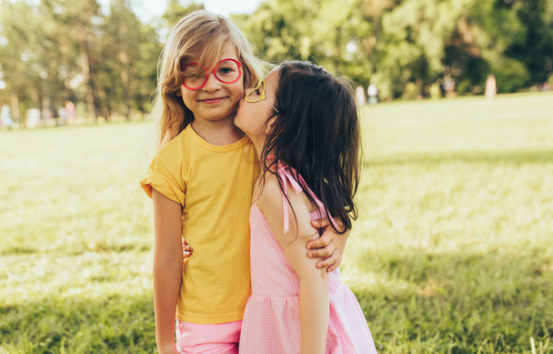 Two positive children shares love and frienship. Two little girls enjoying summertime in the park. Two sisters having fun on sunlight outdoors. Childhood and friendship concept - Photo, image