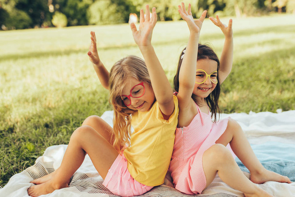Smiling funny children wearing paper glasses with hands up, celebrating the birthday party outdoors, sitting on the blanket. Two little girls having fun in the park. Friends having fun on green grass. - Fotó, kép