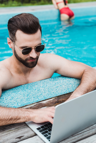 bel homme barbu en lunettes de soleil en utilisant un ordinateur portable dans la piscine
  - Photo, image