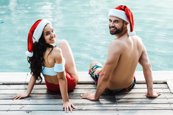 wesoły mężczyzna i kobieta w Santa Claus kapelusze siedzi na drewnianych pokładach w pobliżu basenu  - Zdjęcie, obraz
