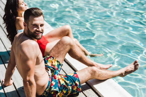 Selektiver Fokus des fröhlichen Mannes, der in die Kamera neben der Frau blickt, während er auf Holzdecks in der Nähe des Schwimmbades sitzt  - Foto, Bild