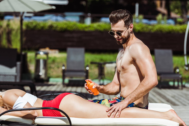szczęśliwy człowiek trzyma butelkę z filtrem przeciwsłonecznym w pobliżu kobiety leżącej na leżaku i patrząc na butelkę w pobliżu człowieka  - Zdjęcie, obraz