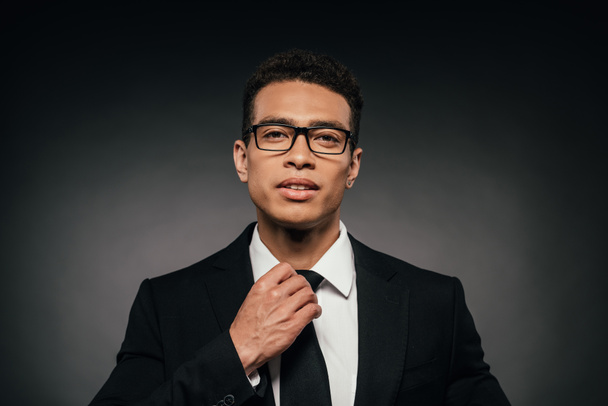handsome african american businessman in glasses and suit touching tie on dark background - Фото, изображение