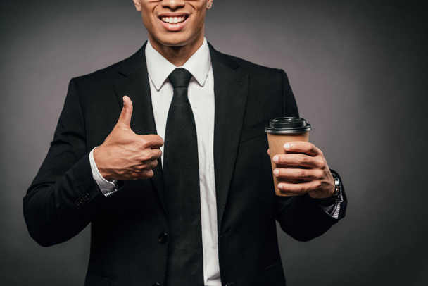 vista ritagliata di sorridente uomo d'affari afro-americano in giacca e cravatta che tiene il caffè per andare e mostrando pollice su sfondo scuro
 - Foto, immagini