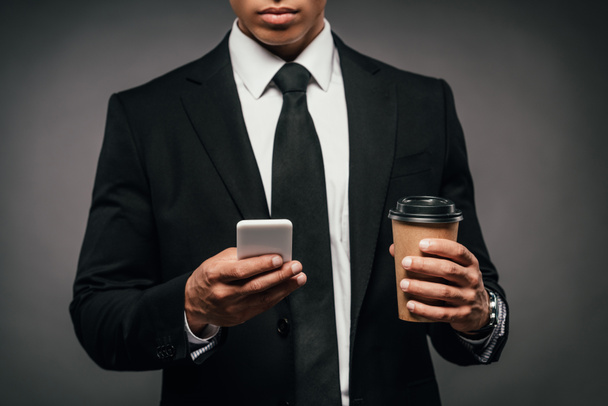 περικομμένη θέα του Αφρικανού Αμερικανού επιχειρηματία που κρατά καφέ για να πάει και smartphone σε σκοτεινό φόντο - Φωτογραφία, εικόνα